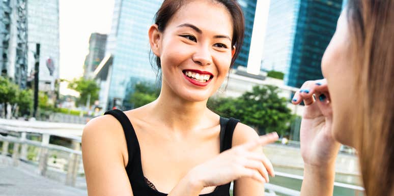 A bright photo of an Asian woman in a black tank top smiling warmly and talking with a friend outdoors near a city waterfront, representing casual charisma, effortless connection, and engaging social conversation.