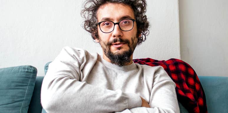 A close-up portrait of a middle-aged man with curly dark hair, glasses, and a beard sitting on a teal couch with his arms tightly crossed, gazing at the camera with a stubborn, distant expression, representing emotional sulking and defiance.