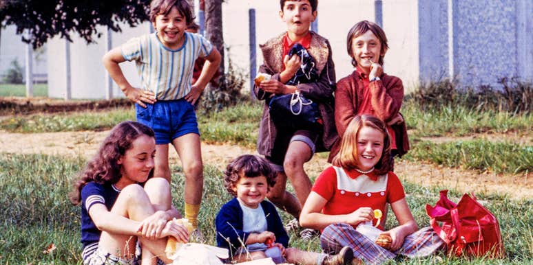 Group of six smiling children from the 1970s sitting and standing on a grassy field, eating snacks.