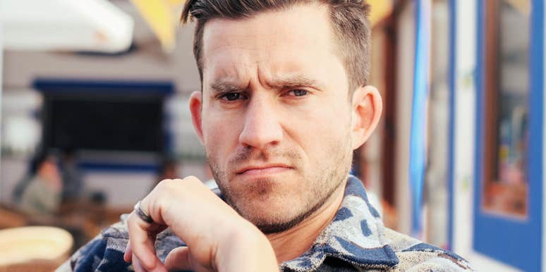 Man with short brown hair and stubble frowning skeptically at the camera with his hand at his chin, seated at an outdoor cafe.