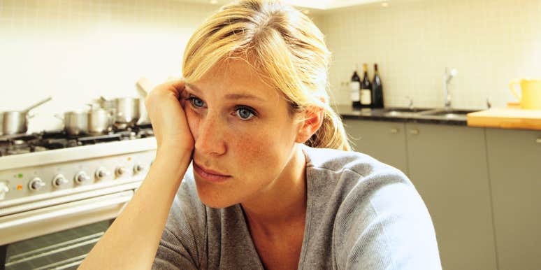 A close-up portrait of a young blonde woman sitting in a modern kitchen, resting her head on her hand with a sad, contemplative look in her blue eyes, representing heartbreak, grief, and life transition.