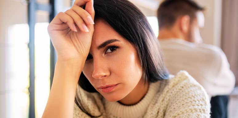 A close-up portrait of a dark-haired woman in a cream sweater resting her head on her hand with a sad, stressed expression, while her husband sits blurred in the background facing away, representing marital strain, hurt feelings, and communication breakdown.