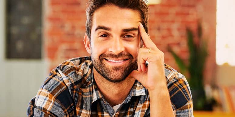 A close-up portrait of a young man with a goatee and thin-framed glasses wearing a burgundy sweater, looking away with a neutral, detached expression in soft sunlight, representing emotional distance, indifference, and introspection.