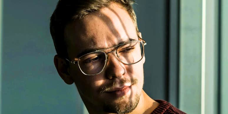 A close-up portrait of a young man with a goatee and thin-framed glasses wearing a burgundy sweater, looking away with a neutral, detached expression in soft sunlight, representing emotional distance, indifference, and introspection.