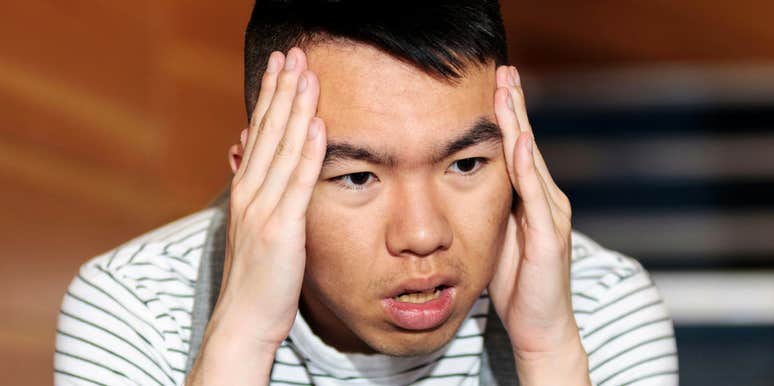 A close-up portrait of an Asian young man in a striped shirt holding both hands to his temples with a stressed, troubled expression, representing relationship anxiety, quiet resignation, and internal conflict.