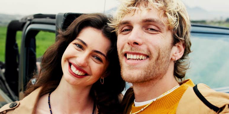 A selfie portrait of a young couple smiling broadly in front of an off-road vehicle outdoors, featuring a dark-haired woman in a beige trench coat leaning against a blonde young man in a yellow sweater, representing genuine happiness, companionship, and shared adventure.