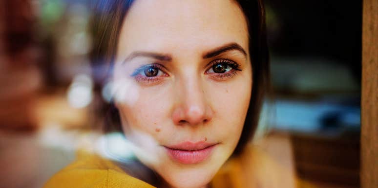Woman with brown eyes, dark hair looking at the camera through a window with soft reflections.