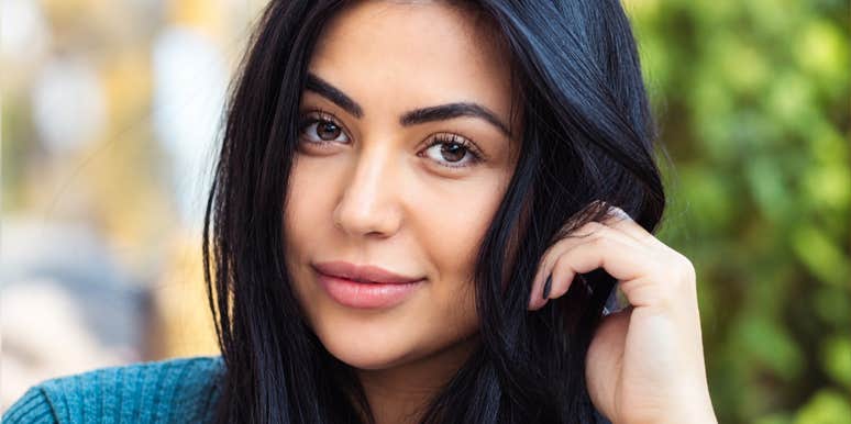 A close-up portrait of a woman with long dark hair and brown eyes looking directly at the camera with a subtle smile while gently touching her hair, representing strong eye contact, magnetic presence, and interpersonal warmth.