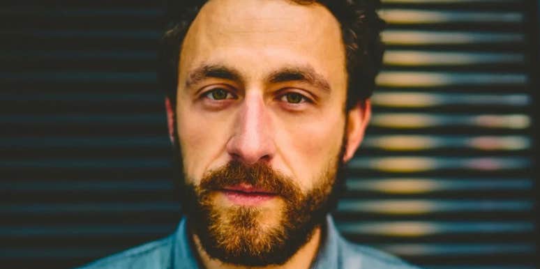 A close-up portrait of a bearded middle-aged man looking directly at the camera with a serious, melancholic expression against a dark background.