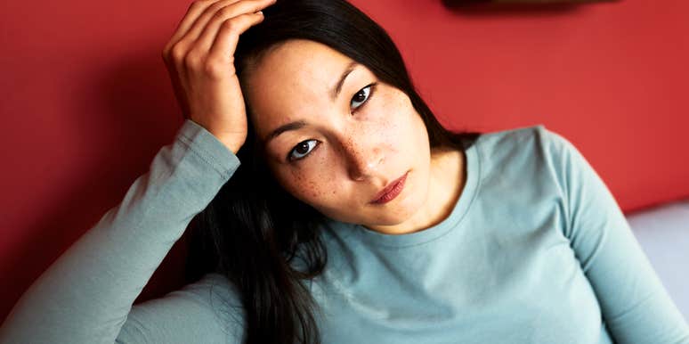 Woman with long dark hair and freckles resting her head on her hand and looking tired and unhappy at the camera against a red wall.