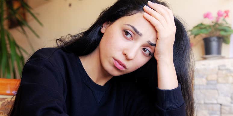 Woman with long dark hair resting her hand on her forehead and looking tired and emotionally drained at the camera in a warmly lit room.