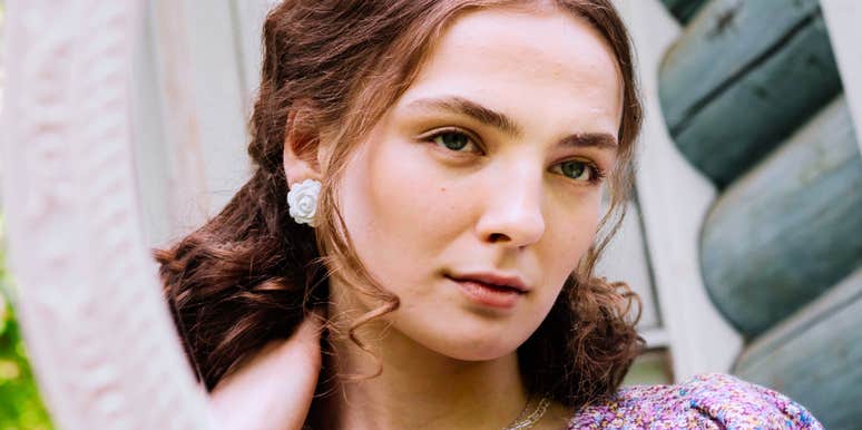 A young woman with soft brown waves and white rose earrings looking calmly into a framed mirror outdoors against a log cabin wall, representing self-reflection, personal value, poised confidence, and boundary setting.