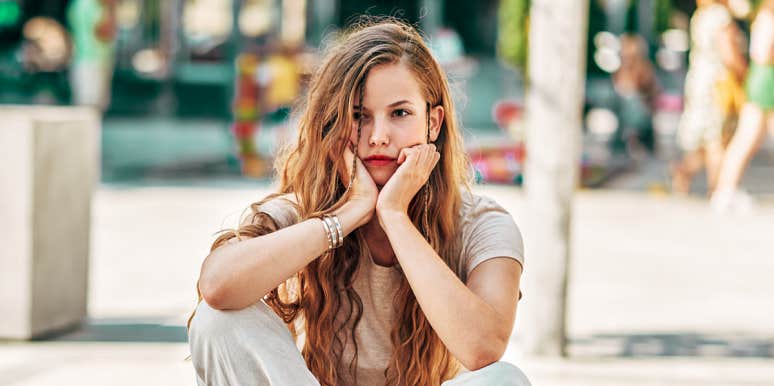 young white woman with long hair looks depressed, head in hands, on city street
