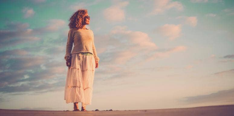 15 Ways Women's Bodies Change As We Age beautiful aging woman with curly brown hair stands on sand in front of vibrant sky