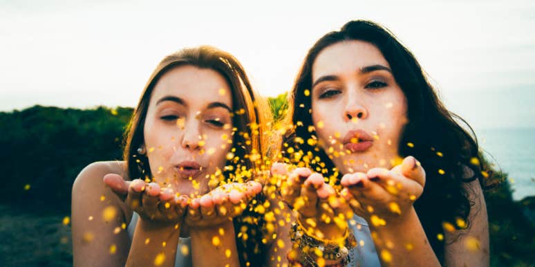 a blonde woman and a brunette woman blow yellow petals at the camera