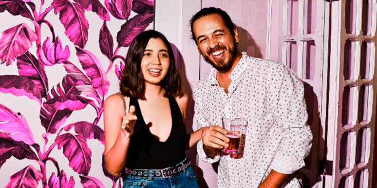 young couple at party against pink floral background, laughing, her hand on his arm
