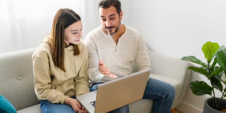 father and teen daughter sitting on couch and talking while looking at laptop