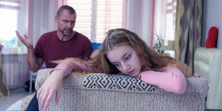 A photograph of a girl reclining on the back of a chair, looking upset as her dad argues with her in the background.