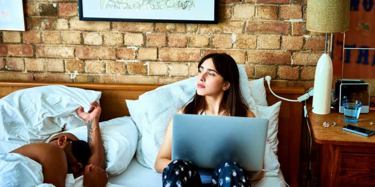 woman looking worried and holding her laptop in bed while man is asleep next to her