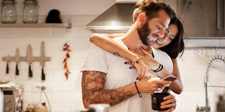 100 Flirty Text Messages To Send To Flirt With A Guy Over Text woman hugging man and looking over his shoulder at smartphone