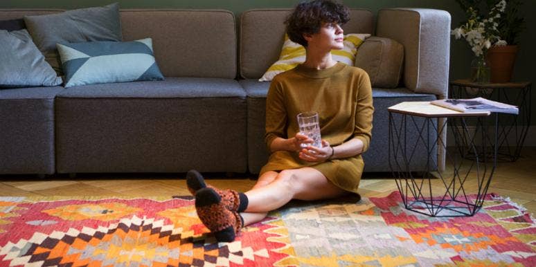 woman with short brown hair wearing a green dress sits on a patterned rug and looks toward a window