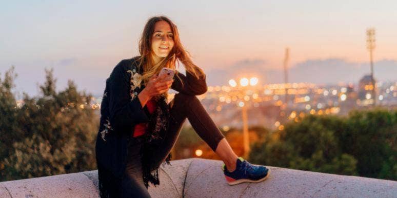 How To Tell If A Girl Likes You Over Text: 18 Signs woman smiling sitting on ledge with phone in her hand
