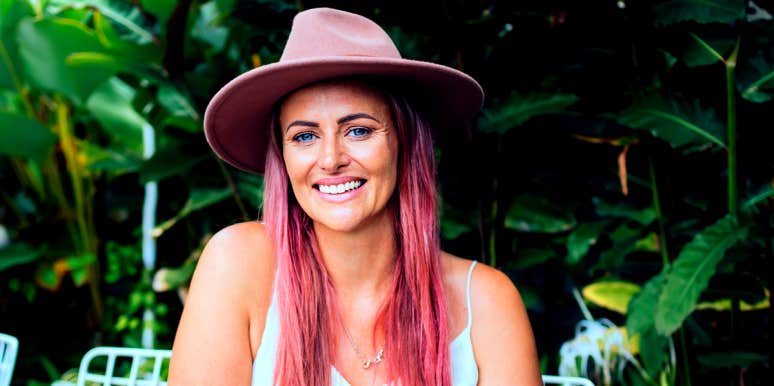 woman in her 30s with pink hair in a pink hat sits in front of greenery