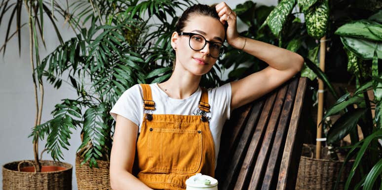 woman in yellow overalls relaxes against a wooden chair by indoor plants