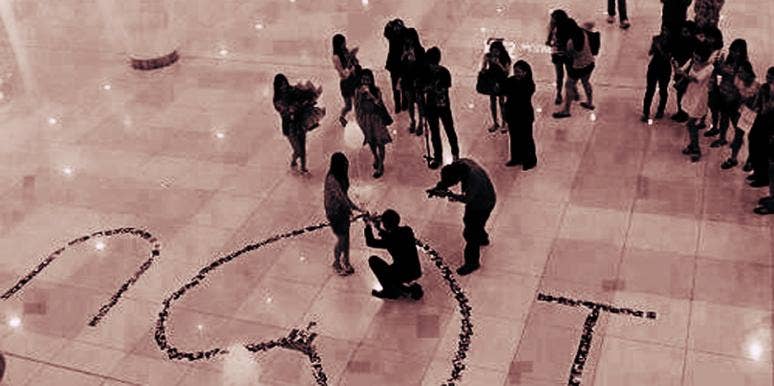 A man gets down on one knee surrounded by rose petals spelling out "I heart you" to propose to his girlfriend that he loves and give her an engagement ring at a New Year's Eve party in front of her family and friends.