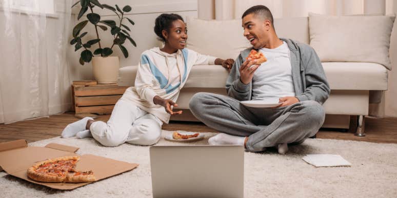 couple sitting on carpet in front of couch eating pizza