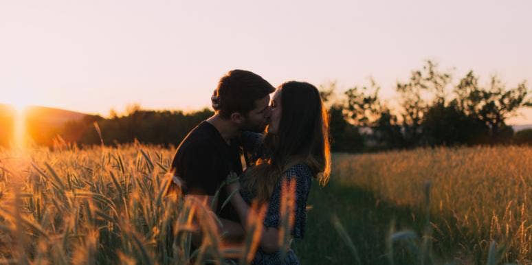 man and woman, each with brown hair, kiss in a field at sunset