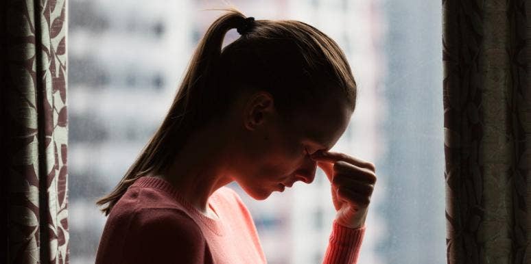 11 Signs Of Trauma Bonding & How To Break The Cycle woman in ponytail pinching the bridge of her nose in front of window