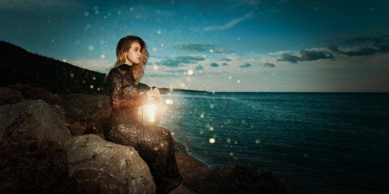 witchy looking woman sits on a rock. at sunset, looking at ocean