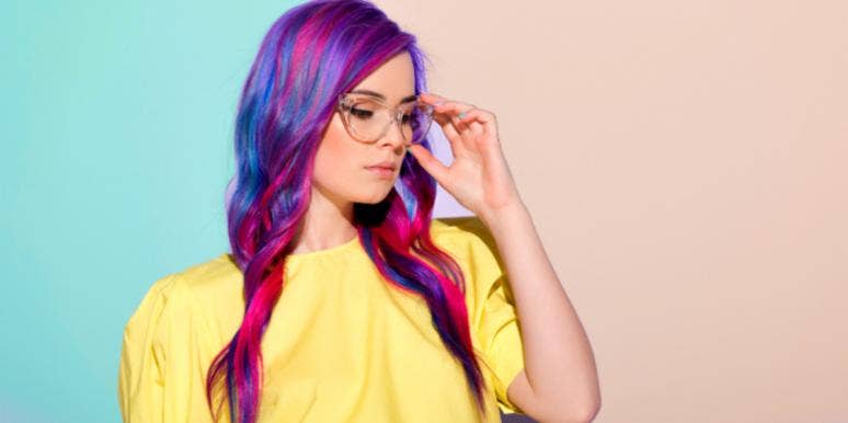 young woman with colorful long hair, wearing a yellow top, lightly touches her glasses, looking away