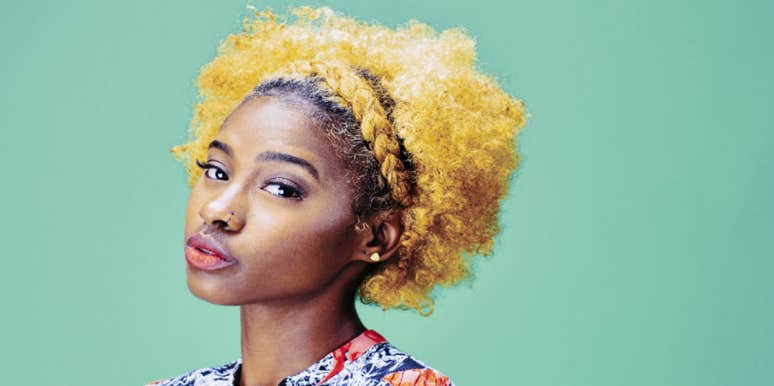 Black woman with blonde hair against green background, looking at camera