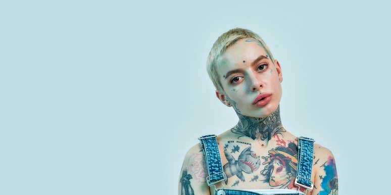  tattooed and pierced white girl wearing denim overall standing and looking into a camera on a light blue color background