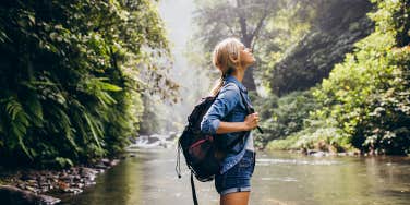 Side view shot relaxed young woman with backpack standing by the stream. Female hiking by the creek.