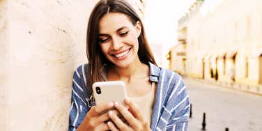 Woman leaning on wall smiling big, texting.