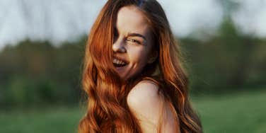 A young woman smiles and looks into the camera with her long, red, wavy, shiny hair in a park with green grass