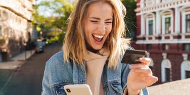 Beautiful cheerful young blonde woman wearing denim jacket walking in the city, using mobile phone, showing plastic credit card