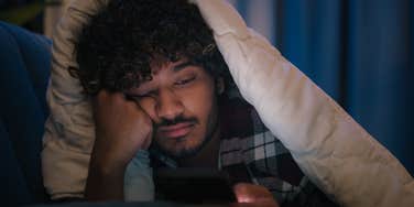 Sad man laying in bed scrolling on his phone.