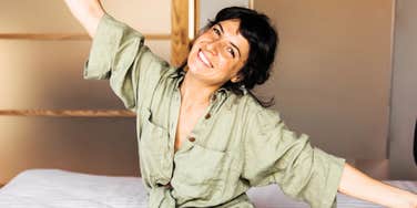 Happy young woman sitting on bed, smiling and spreading her arms in a bright minimalist room, illustrating the joyful habits of naturally minimalist people and intentional simple living.