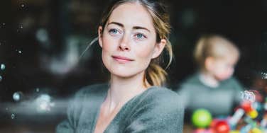 A calm woman gazes out the window while her child plays behind her, symbolizing self-care, emotional boundaries, and the power of learning to put herself first without guilt.