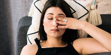 Exhausted woman rubbing her eyes on the couch, illustrating the quiet guilt people feel for resting due to habits learned in childhood.