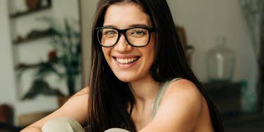 Smiling woman wearing glasses sitting comfortably indoors, appearing relaxed, confident, and emotionally grounded.