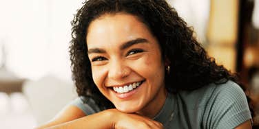 Smiling person with curly hair resting their arms on a table, appearing relaxed, confident, and genuinely at ease in a softly lit space.
