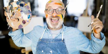 Ecstatic mature artist holding a palette and paintbrushes showing the art of starting over and the wild choices that completely rewrite your life.