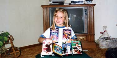 Child smiling while holding VHS tapes in a living room with a box television, capturing a nostalgic nineties family moment centered around shared movies and time together.