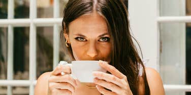 Woman drinking from a cup while standing near a window in natural light.