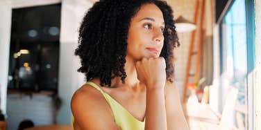 Adult woman sitting indoors in natural light, resting her chin on her hand and looking thoughtfully out a window.